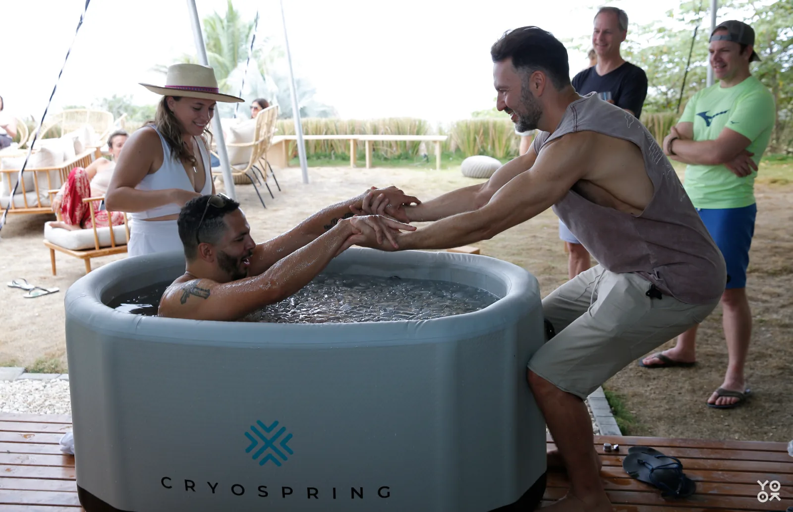 A cold plunge session on a wooden deck, with one man in an ice bath holding hands with another